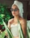 A person wearing a towel turban and fashionable sunglasses is surrounded by lush green plants. The setting is serene, with soft lighting enhancing their contemplative expression. The person has a spa-like appearance, adding a sense of relaxation to the atmosphere.