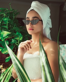 A person wearing a towel turban and fashionable sunglasses is surrounded by lush green plants. The setting is serene, with soft lighting enhancing their contemplative expression. The person has a spa-like appearance, adding a sense of relaxation to the atmosphere.