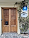 A wooden double door with intricate lattice patterns, flanked by a leafy plant with purple flowers. Above the door are signs bearing the words 'Hotel America' and a blue hotel badge indicating 'H Ciudad.' The stone path leading up to the entrance adds a rustic charm.