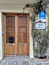 A wooden double door with intricate lattice patterns, flanked by a leafy plant with purple flowers. Above the door are signs bearing the words 'Hotel America' and a blue hotel badge indicating 'H Ciudad.' The stone path leading up to the entrance adds a rustic charm.