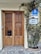 A wooden double door with intricate lattice patterns, flanked by a leafy plant with purple flowers. Above the door are signs bearing the words 'Hotel America' and a blue hotel badge indicating 'H Ciudad.' The stone path leading up to the entrance adds a rustic charm.