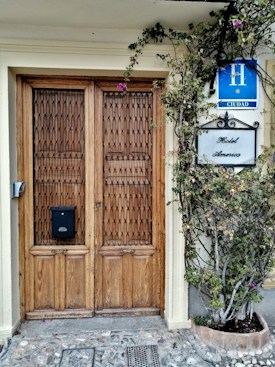 A wooden double door with intricate lattice patterns, flanked by a leafy plant with purple flowers. Above the door are signs bearing the words 'Hotel America' and a blue hotel badge indicating 'H Ciudad.' The stone path leading up to the entrance adds a rustic charm.