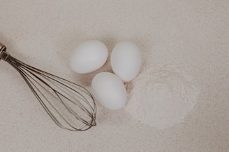 Three white eggs are placed on a light-colored surface next to a mound of flour. A metal whisk is positioned to the left of the eggs and flour.