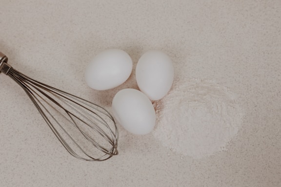 Three white eggs are placed on a light-colored surface next to a mound of flour. A metal whisk is positioned to the left of the eggs and flour.
