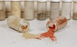 Two spice jars are lying on their sides on a countertop, spilling powders onto the surface. One jar contains a red spice, possibly paprika, while the other has a brownish spice, possibly cumin. Several similar jars are blurred in the background.