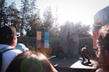 A group of performers in costumes are engaging in a choreographed action scene in front of a large stone backdrop. Banners hang beside the stone, and spectators watch the event intently from the foreground. The scene is set outdoors with tall trees in the background and bright sunlight creating lens flares.