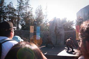 A group of performers in costumes are engaging in a choreographed action scene in front of a large stone backdrop. Banners hang beside the stone, and spectators watch the event intently from the foreground. The scene is set outdoors with tall trees in the background and bright sunlight creating lens flares.