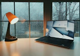 A cozy desk setup with a laptop, notebook, and a cup of coffee beside a window showing a rainy day.
