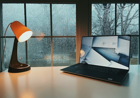 A cozy workspace with a laptop, notebook, and a cup of tea beside a window showing a soft rainy day.