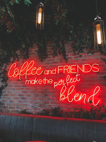 A neon sign with the phrase 'Coffee and FRIENDS make the perfect blend' is mounted on a wall with red bricks and adorned with green vines. Hanging pendant lights are also visible in the scene. Below the sign is a cushioned bench seating area.