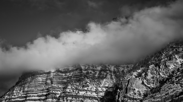 A striking black and white photograph of a mountain partially shrouded in soft, low-hanging clouds. The mountain features distinct, horizontal rock strata, and its slopes are dotted with patches of snow. Dense, rugged vegetation clings to the steep sides.