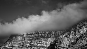 A striking black and white photograph of a mountain partially shrouded in soft, low-hanging clouds. The mountain features distinct, horizontal rock strata, and its slopes are dotted with patches of snow. Dense, rugged vegetation clings to the steep sides.
