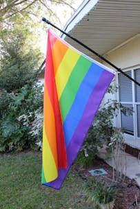 A rainbow flag is mounted on the side of a house. The flag displays vibrant colors and is positioned in front of a window with blinds. Lush greenery, including bushes and a well-maintained lawn, surrounds the area. The house's exterior features light-colored walls and an overhanging roof.