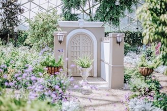 A picturesque garden features a decorative archway with an elegant white door in the center, flanked by two light fixtures. The scene is filled with greenery, including trees and various plants, and colorful flowers, particularly purple ones, add a vibrant touch to the tranquil setting. A potted plant is positioned in front of the door, enhancing the serene garden atmosphere. A dome-like glass structure above suggests a conservatory setting.