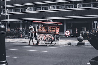 A street vendor pushes a food cart with signs displaying 'Nasi Goreng' and 'Soto Ayam'. The scene takes place on a quiet urban street with a large building in the background. The vendor is wearing casual clothing and the cart is equipped with two large wheels. Some plants and a no-parking sign are visible nearby.
