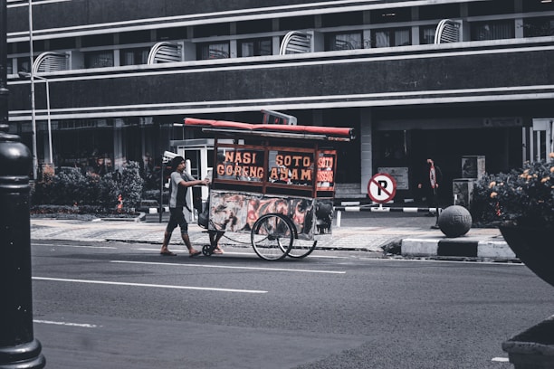 A street vendor pushes a food cart with signs displaying 'Nasi Goreng' and 'Soto Ayam'. The scene takes place on a quiet urban street with a large building in the background. The vendor is wearing casual clothing and the cart is equipped with two large wheels. Some plants and a no-parking sign are visible nearby.