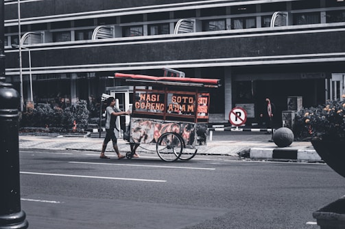 A street vendor pushes a food cart with signs displaying 'Nasi Goreng' and 'Soto Ayam'. The scene takes place on a quiet urban street with a large building in the background. The vendor is wearing casual clothing and the cart is equipped with two large wheels. Some plants and a no-parking sign are visible nearby.
