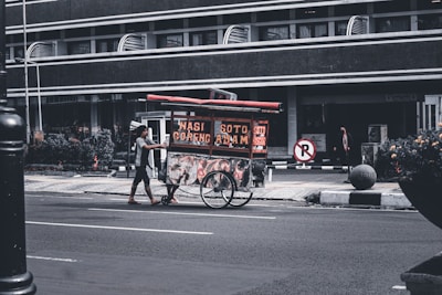 A street vendor pushes a food cart with signs displaying 'Nasi Goreng' and 'Soto Ayam'. The scene takes place on a quiet urban street with a large building in the background. The vendor is wearing casual clothing and the cart is equipped with two large wheels. Some plants and a no-parking sign are visible nearby.