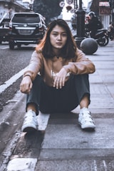 A young person is sitting on the sidewalk of an urban street. They are wearing casual clothing, including a brown hoodie and white sneakers. The background shows parked cars, a motorcycle, street signs, and several pedestrians.