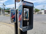 A public payphone with a blue handset and coin slot is mounted on a sidewalk pole on a street corner. The phone booth features colorful advertisements promoting savings. Nearby, a second smaller box is attached to the pole, also decorated with similar advertisements. The background shows a quiet intersection with visible traffic lights and a gas station across the street.
