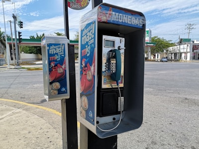 A public payphone with a blue handset and coin slot is mounted on a sidewalk pole on a street corner. The phone booth features colorful advertisements promoting savings. Nearby, a second smaller box is attached to the pole, also decorated with similar advertisements. The background shows a quiet intersection with visible traffic lights and a gas station across the street.