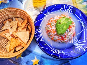 A woven basket filled with crispy tortilla chips sits next to a vibrant blue plate. The plate holds a neatly formed ceviche topped with a slice of avocado, showcasing a mix of diced seafood, vegetables, and herbs. Beneath them, a colorful table surface with star patterns adds a decorative background.