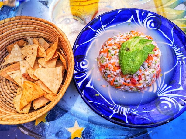 A woven basket filled with crispy tortilla chips sits next to a vibrant blue plate. The plate holds a neatly formed ceviche topped with a slice of avocado, showcasing a mix of diced seafood, vegetables, and herbs. Beneath them, a colorful table surface with star patterns adds a decorative background.