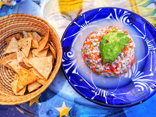 A woven basket filled with crispy tortilla chips sits next to a vibrant blue plate. The plate holds a neatly formed ceviche topped with a slice of avocado, showcasing a mix of diced seafood, vegetables, and herbs. Beneath them, a colorful table surface with star patterns adds a decorative background.