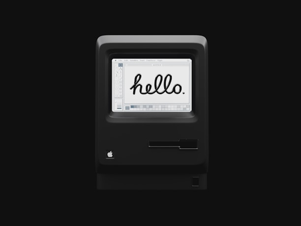A vintage computer with a monochrome screen displaying the word 'hello' in bold, black script against a white background. The computer casing is black with a classic design, featuring an Apple logo. The screen interface shows early graphic design software elements.