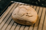 A freshly baked loaf of bread with a rustic, crusty exterior rests on a cooling rack. The bread appears golden brown and well-risen, with a few cracks and a rough texture on the surface.