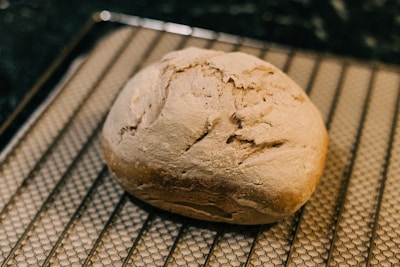 A freshly baked loaf of bread with a rustic, crusty exterior rests on a cooling rack. The bread appears golden brown and well-risen, with a few cracks and a rough texture on the surface.