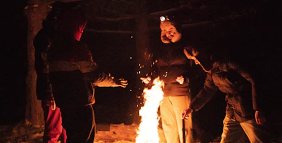 Group of friends in The North Face parkas laughing around a campfire in a snowy forest.
