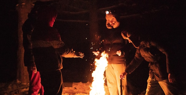 A group of guys wearing rowdy.co apparel, gathered around a campfire in the woods at dusk.