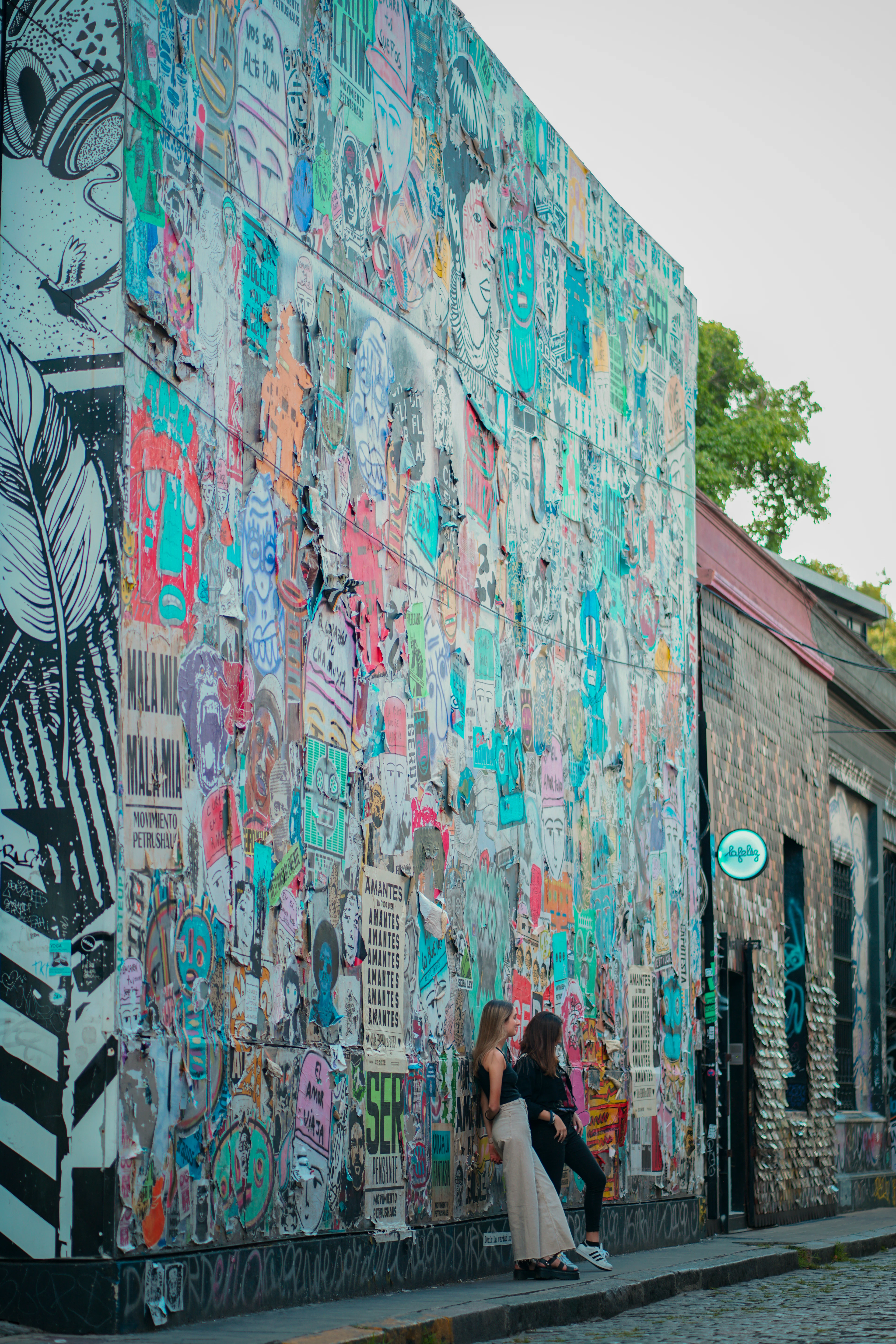 Two women lean against a wall covered in colorful street art stickers on a cobblestone street.