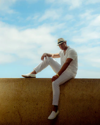 man in white long sleeve shirt and white pants sitting on brown concrete wall during daytime