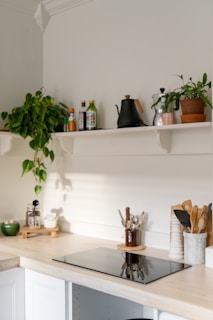 A minimalist kitchen corner featuring a light wooden countertop with an induction cooktop. Above the countertop, there are white shelves holding various items including potted plants, a black kettle, condiments, and jars. Kitchen utensils are neatly organized in containers beside the cooktop, and the area is well-lit with natural daylight streaming in.
