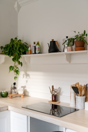 A minimalist kitchen corner featuring a light wooden countertop with an induction cooktop. Above the countertop, there are white shelves holding various items including potted plants, a black kettle, condiments, and jars. Kitchen utensils are neatly organized in containers beside the cooktop, and the area is well-lit with natural daylight streaming in.