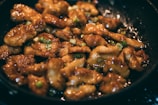 Pieces of golden-brown chicken coated in a glossy sauce fill a frying pan, garnished with chopped green onions.