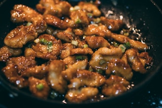 Pieces of golden-brown chicken coated in a glossy sauce fill a frying pan, garnished with chopped green onions.