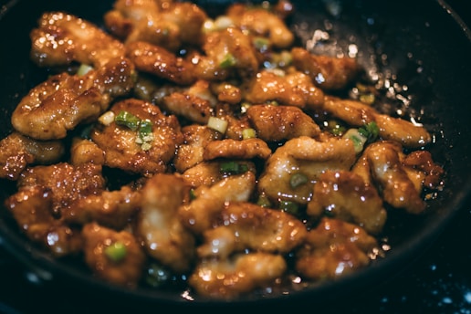 Pieces of golden-brown chicken coated in a glossy sauce fill a frying pan, garnished with chopped green onions.