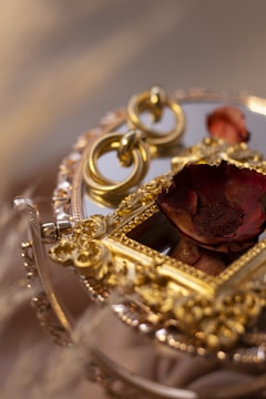 An ornate, baroque-style gilded frame set against a reflective surface, accompanied by a dried red flower. The intricate gold detailing includes floral patterns, adding to the luxurious and vintage aesthetic.