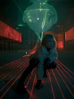 A person crouches on a path illuminated by red laser beams that crisscross the scene. The background features a large, transparent inflatable structure under green lighting, creating a futuristic and immersive environment.