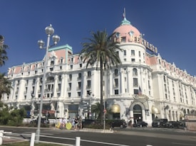 A grand, historic building with white walls and a pink dome roof. The building has intricate architectural details and a sign with the name 'Le Negresco'. In front of the building, there are several parked cars, a tall palm tree, and a street with a few people walking.