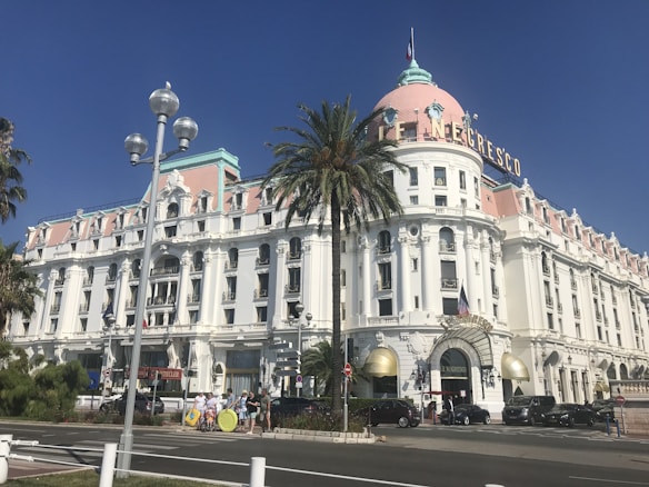 A grand, historic building with white walls and a pink dome roof. The building has intricate architectural details and a sign with the name 'Le Negresco'. In front of the building, there are several parked cars, a tall palm tree, and a street with a few people walking.