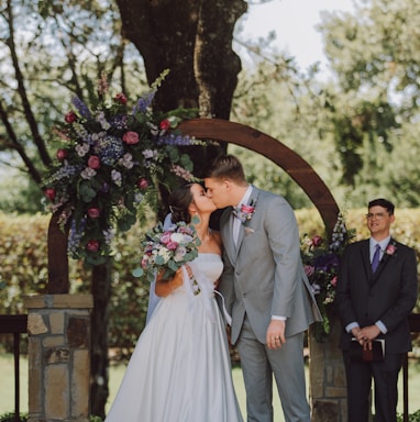 man in black suit and woman in white wedding dress kissing under green tree during daytime