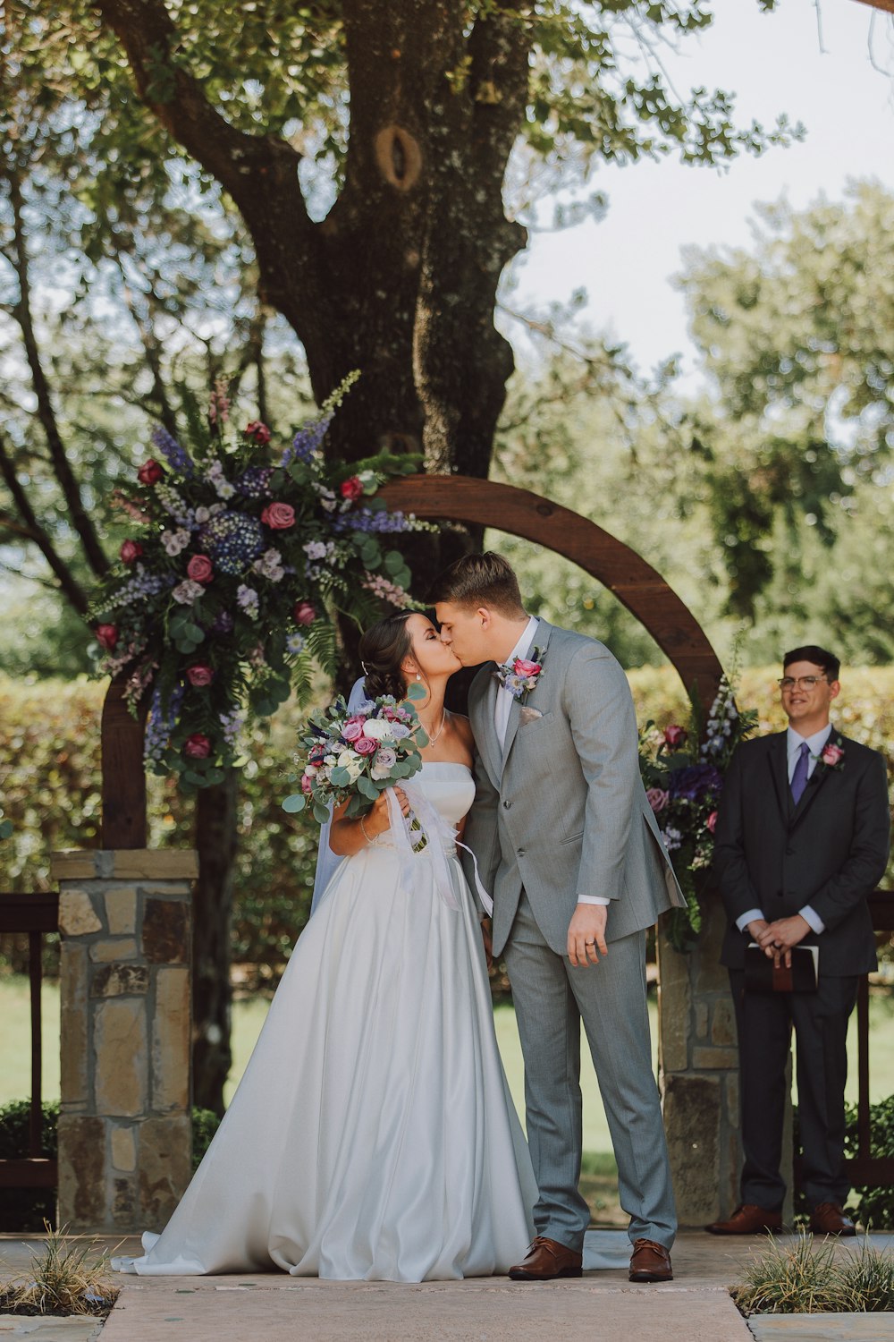 man in black suit and woman in white wedding dress kissing under green tree during daytime