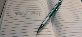An open lined notebook with Japanese writing is placed on a surface. A pen, designed with a silver barrel and a black grip, lies diagonally across the pages.