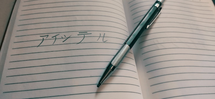An open lined notebook with Japanese writing is placed on a surface. A pen, designed with a silver barrel and a black grip, lies diagonally across the pages.