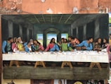A painted recreation of 'The Last Supper' featuring Jesus at the center and surrounded by disciples in a room with dark walls and a wooden ceiling. The disciples are engaged in animated discussion, and a long table stretches across the image, covered with a white tablecloth and various dishes.