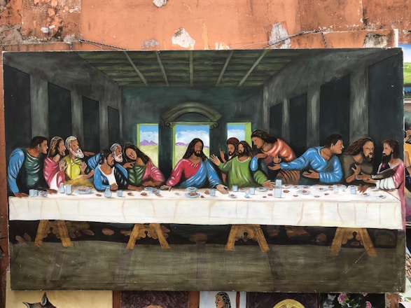 A painted recreation of 'The Last Supper' featuring Jesus at the center and surrounded by disciples in a room with dark walls and a wooden ceiling. The disciples are engaged in animated discussion, and a long table stretches across the image, covered with a white tablecloth and various dishes.